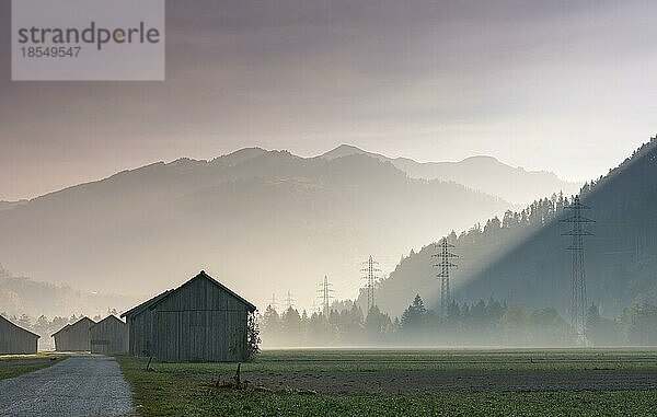 Blick auf das Prättigautal bei Klosters in der Schweiz an einem frühen Morgen mit Feldern und Holzscheunen  Stromleitungen und Bergen in der Silhouette im Spätherbst