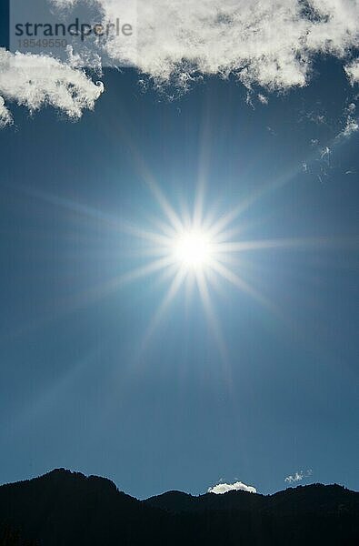 Sonne scheint hell in einer Sternform in den hellblauen Himmel mit ein paar weißen Wolken und einem Berg Silhouette unten als vertikalen Hintergrund