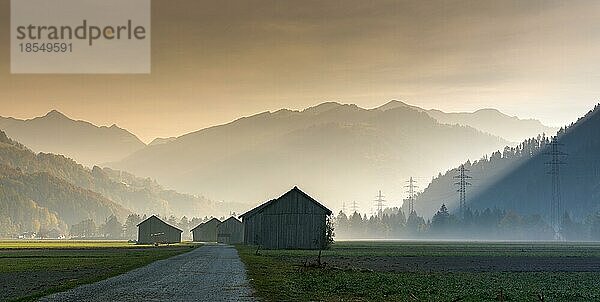 Blick auf das Prättigautal bei Klosters in der Schweiz an einem frühen Morgen mit Feldern und Holzscheunen  Stromleitungen und Bergen in der Silhouette im Spätherbst