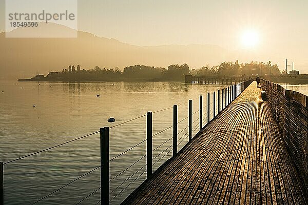 Langer Holzsteg und Promenade über dem Zürichsee bei Rapperswil im goldenen Abendlicht mit Bergsilhouette im Hintergrund