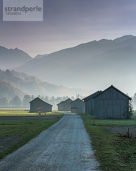 Blick auf das Prättigautal bei Klosters in der Schweiz an einem frühen Morgen mit Feldern  Holzscheunen und Bergen in der Silhouette im Spätherbst