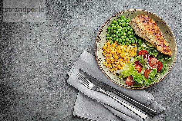Gebackene Hähnchenbrust auf Teller mit frischem Salat  grünen Erbsen und Mais  grauer Steinhintergrund  Draufsicht. Gesunde FitnessMahlzeit mit Hähnchenfilet  ausgewogen in Proteinen und Kohlenhydraten. Overhead  Kopierbereich