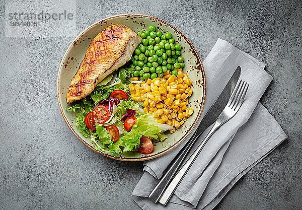 Gebackene Hähnchenbrust auf Teller mit frischem Salat  grünen Erbsen und Mais  grauer Steinhintergrund  Draufsicht. Gesunde FitnessMahlzeit mit Hähnchenfilet  ausgewogen in Proteinen und Kohlenhydraten. Diät und Ernährung