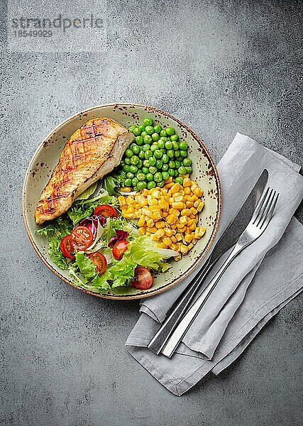 Gebackene Hähnchenbrust auf Teller mit frischem Salat  grünen Erbsen und Mais  grauer Steinhintergrund  Draufsicht. Gesunde FitnessMahlzeit mit Hähnchenfilet  ausgewogen in Proteinen und Kohlenhydraten. Diät und Ernährung