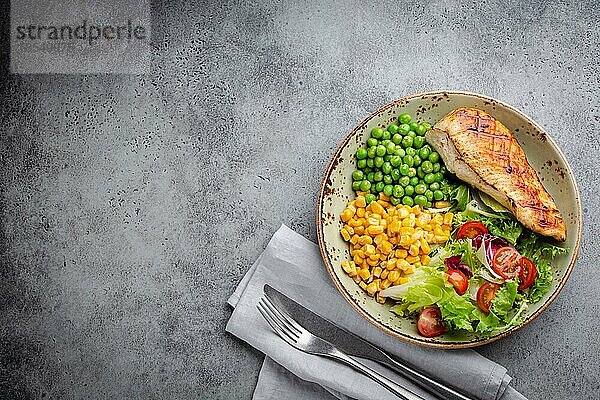 Gebackene Hähnchenbrust auf Teller mit frischem Salat  grünen Erbsen und Mais  grauer Steinhintergrund  Draufsicht. Gesunde FitnessMahlzeit mit Hähnchenfilet  ausgewogen in Proteinen und Kohlenhydraten. Overhead  Kopierbereich