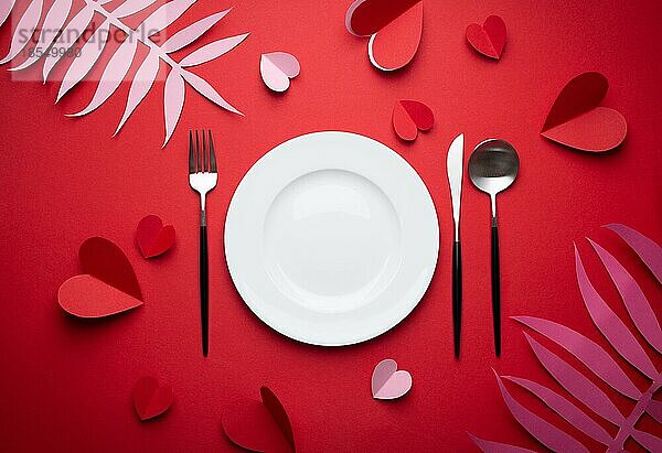 Romantisches Abendessen im Restaurant am Valentinstag  Papierkunst. Weiß leeren Teller  Löffel  Gabel  Messer auf rotem Hintergrund mit Papier Herzen und tropischen Blättern. Valentinstag Datum Konzept Handwerk Origami