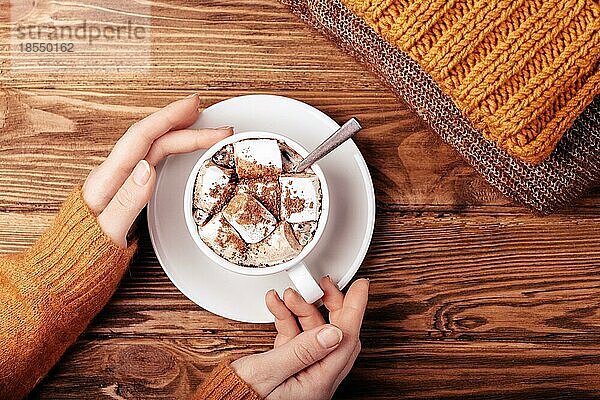 Gemütliche Winter flach legen  weibliche Hände halten Tasse mit heißer Schokolade oder Kakao mit Marshmallow und Stapel von warmen gestrickte Kleidung auf Holz rustikalen Hintergrund. Gemütlich und weich Winter oder Herbst Konzept