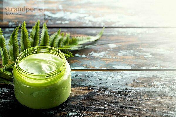 Kosmetische Creme in einem Behälter auf einem hölzernen Hintergrund mit grünem Blatt  Naturkosmetik  Bio Hautpflegeprodukt Konzept. Hausgemachte Creme für Gesicht und Körperpflege  Hintergrund mit Kopie Raum Draufsicht