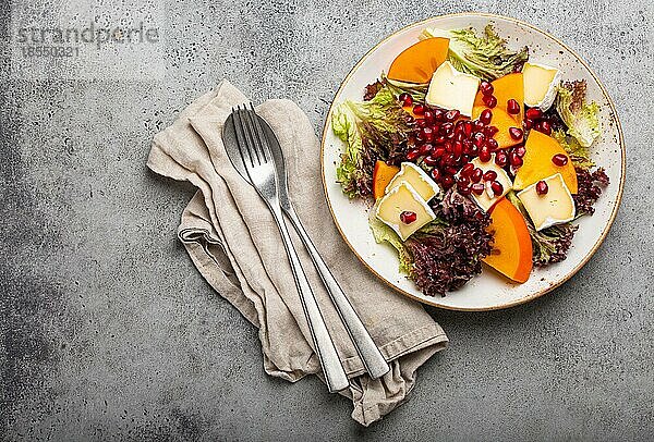 Draufsicht flach legen Kaki-Salat mit Brie-Käse  frischer Salat Blätter  Granatapfelkerne auf weißem Teller steingrauem Hintergrund  Obstsalat der Saison als Vorspeise  vegetarische gesunde Lebensmittel Kopie Raum