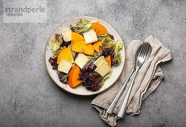 Draufsicht flach legen Kaki-Salat mit Brie-Käse und frischem Salat Blätter auf weißem Teller und Stein grauem Hintergrund  Obstsalat der Saison als Vorspeise  vegetarische gesunde Ernährung