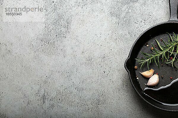 Schwarzes Gusseisen Bratpfanne mit Zutaten für das Kochen: Öl  frischer grüner Rosmarin  rohe Knoblauchzehen und bunte Pfefferkörner auf grauem Stein Hintergrund flach legen Draufsicht mit Platz für Text