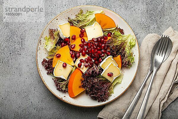Draufsicht flach legen Kaki-Salat mit Brie-Käse  frische Salatblätter  Granatapfelkerne auf weißem Teller und steingrauem Hintergrund  Obstsalat der Saison als Vorspeise  vegetarische gesunde Ernährung