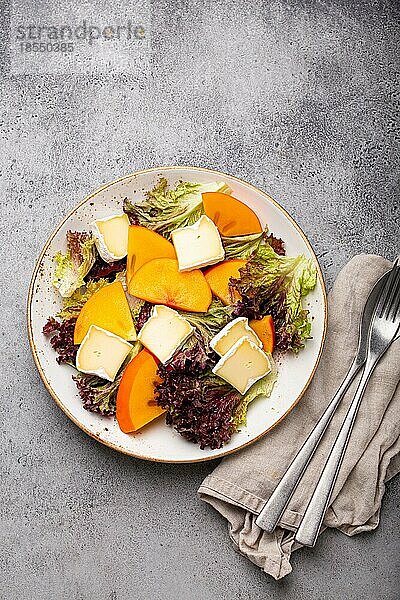 Draufsicht flach legen Kaki-Salat mit Brie-Käse und frischem Salat Blätter auf weißem Teller und Stein grauem Hintergrund  Obstsalat der Saison als Vorspeise  vegetarische gesunde Ernährung