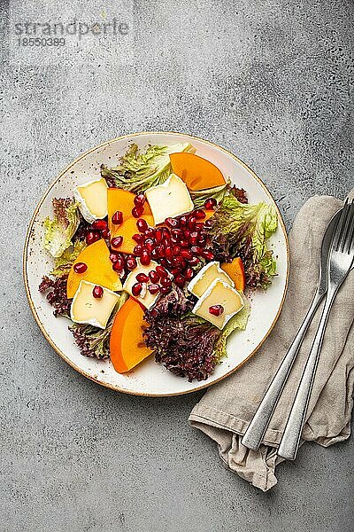 Draufsicht flach legen Kaki-Salat mit Brie-Käse  frische Salatblätter  Granatapfelkerne auf weißem Teller und steingrauem Hintergrund  Obstsalat der Saison als Vorspeise  vegetarische gesunde Ernährung