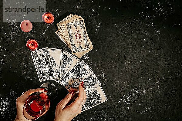 Esoterische Komposition Draufsicht mit weiblichen Händen einer Wahrsagerin mit Glas Rotwein und Zigarette  die Tarotkarten auf einem schwarzen magischen Tisch mit roten Kerzen liest  Okkultismus und Prognosen  Kopierraum