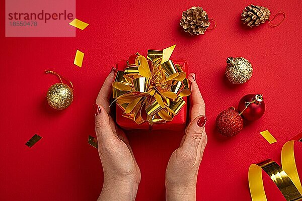 Weibliche Hände mit roten Nägeln halten Weihnachtsgeschenk-Box in rotem Papier mit goldenen Band und Bogen eingewickelt  rote Weihnachten funkelnden runden Kugeln Dekorationen auf rotem Papier Hintergrund Draufsicht flach legen