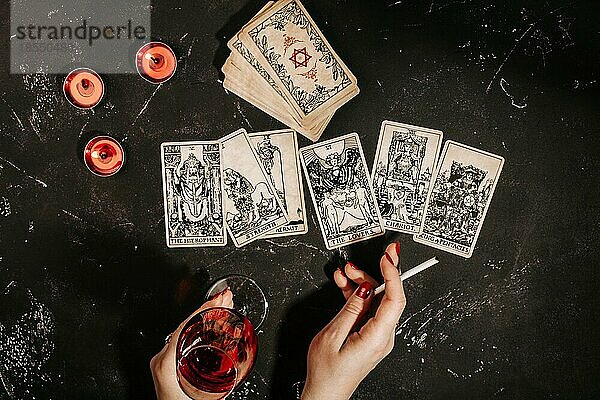 Esoteric Zusammensetzung Draufsicht mit weiblichen Händen der Wahrsagerin mit Glas Rotwein und Zigarette lesen Tarot-Karten auf schwarze Magie Tabelle mit roten brennenden Kerzen  okkulte und Prognose Konzept