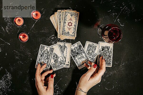 Esoteric Zusammensetzung Draufsicht mit weiblichen Händen der Wahrsagerin mit Glas Rotwein Lesen von Tarot-Karten auf schwarze Magie Hintergrund Tabelle mit roten brennenden Kerzen  okkulte und Prognose Konzept