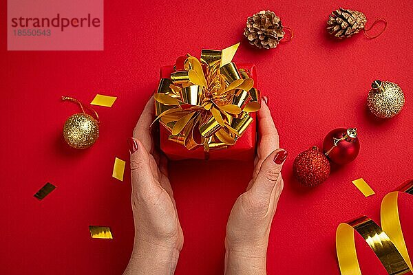 Weibliche Hände mit roten Nägeln halten Weihnachtsgeschenk-Box in rotem Papier mit goldenen Band und Bogen eingewickelt  rote Weihnachten funkelnden runden Kugeln Dekorationen auf rotem Papier Hintergrund Draufsicht flach legen