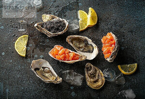Meeresfrüchte  verschiedene Vorspeisen und Häppchen: frische offene Austern  schwarzer Kaviar  Lachstatar  serviert mit Zitronenspalten und Eiswürfeln  Draufsicht  flach liegend  auf blauem Betonsteinhintergrund