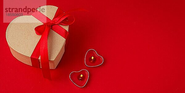 Schöne minimale eingewickelt Geschenk-Box herzförmig mit roter Schleife Band auf rotem Hintergrund und zwei beleuchtete Kerzen Draufsicht flach legen  Geschenk für Saint Valentine Tag  Liebe und Romantik  Platz für Text