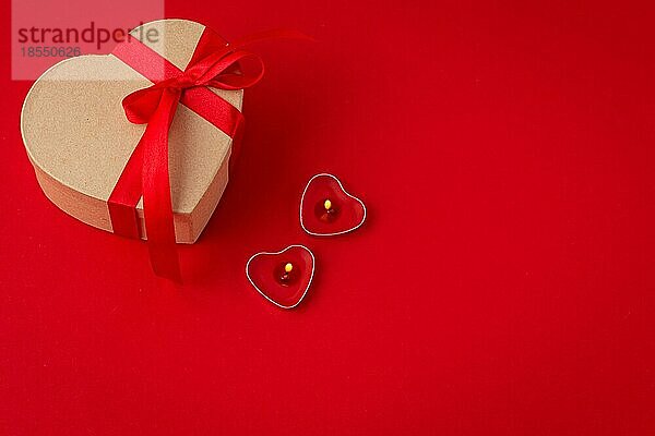 Schöne minimale eingewickelt Geschenk-Box herzförmig mit roter Schleife Band auf rotem Hintergrund und zwei beleuchtete Kerzen Draufsicht flach legen  Geschenk für Saint Valentine Tag  Liebe und Romantik  Platz für Text