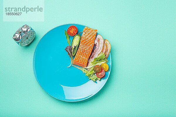 Intermittierendes Fasten niedrige Kohlenhydrate hohe Fette Diät-Konzept flach legen  gesunde Lebensmittel Lachs Fisch  Speck Fleisch  Gemüse und Salat auf blauem Teller und Wecker auf blauem Hintergrund Ansicht von oben