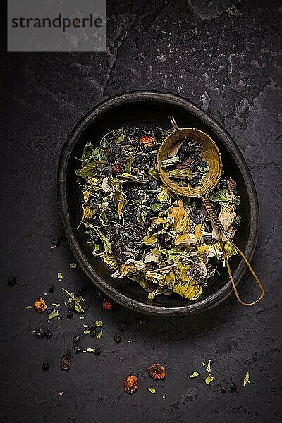 Teemischung  getrocknete Bio Teekräuter und Früchte in Schale mit Teesieb auf schwarzem Hintergrund