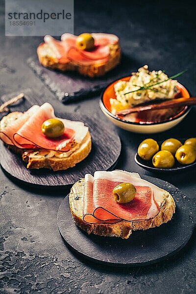 Geröstetes Brot oder offenes Sandwich mit Knoblauchbutteraufstrich und Serano Schinken und Oliven auf dunklem Hintergrund