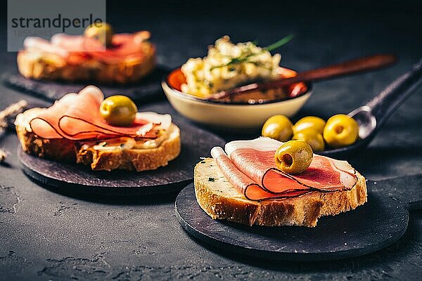 Geröstetes Brot oder offenes Sandwich mit Knoblauchbutteraufstrich und Serano Schinken und Oliven auf dunklem Hintergrund