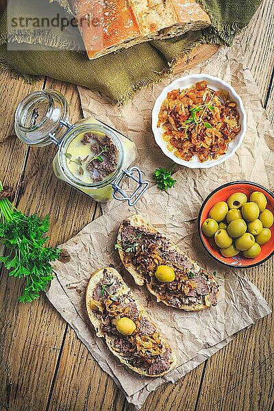 Frische hausgemachte Hühnerleberpastete auf Ciabatta Brot mit gerösteten Zwiebeln und Oliven auf Holztisch