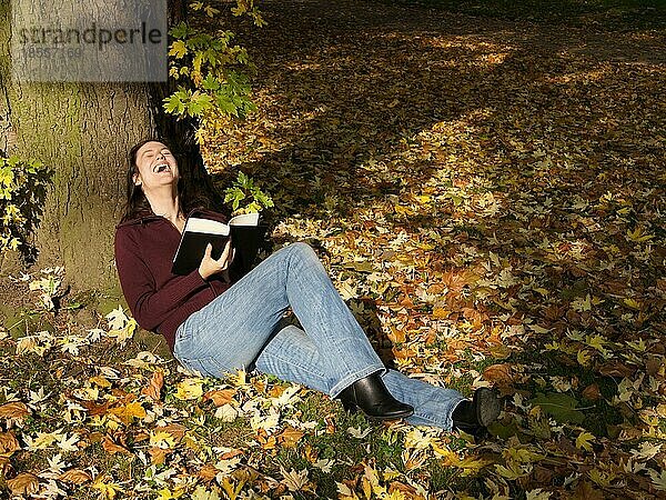 Junge Frau entspannt mit einem Buch in herbstlicher Szene und lacht laut