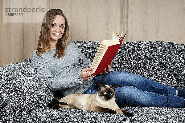 Junge Frau entspannt sich mit einem Buch und ihrer Katze auf der Couch