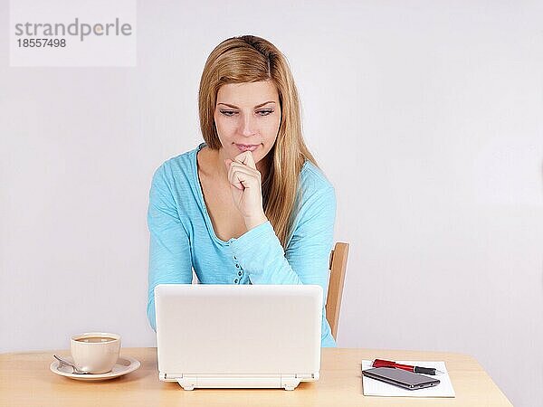 Junge Frau sitzt am Schreibtisch im Home Office mit Laptop und Kaffee