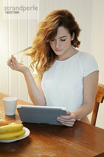 Junge Frau benutzt einen Tablet-Computer  während sie zu Hause am Küchentisch Kaffee trinkt