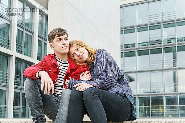 Junges zärtliches heterosexuelles Paar sitzt auf Stufen vor einem modernen Gebäude mit Glasfassade