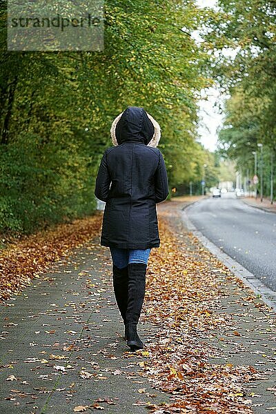 Rückansicht einer nicht erkennbaren Frau in Wintermantel und Stiefeln  die im Herbst die Straße entlanggeht