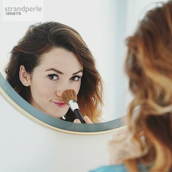 Junge Frau pudert sich die Nase mit einem Schminkpinsel  Badezimmerspiegel  Porträt mit selektivem Fokus