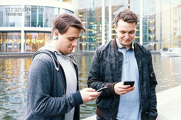 Zwei asoziale handyabhängige männliche Teenager stehen zusammen und benutzen ein Smartphone  Technologiekonzept  städtische Uferlage in Berlin