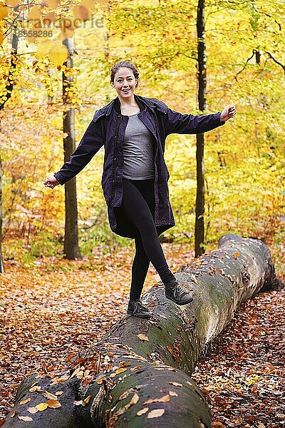 Glückliche  unbekümmerte junge Frau  die im Wald auf einem Baumstamm balanciert - aktiver Lebensstil in der Herbstzeit