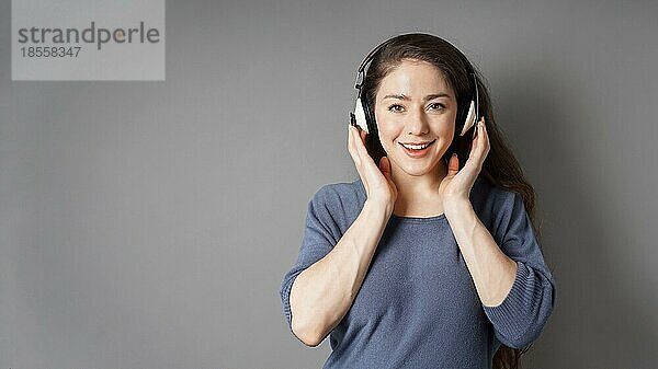 Glücklich lächelnde junge Frau  die mit drahtlosen Kopfhörern Musik hört - grauer Hintergrund mit Kopierraum
