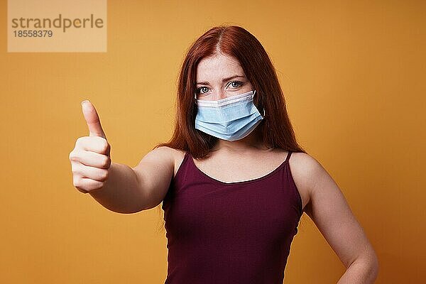 Junge Frau mit medizinischer Gesichtsmaske  die Mund und Nase korrekt bedeckt und den Daumen nach oben streckt - Schutz vor dem Corona-Virus - Studioporträt auf orangefarbenem Hintergrund mit Kopierraum