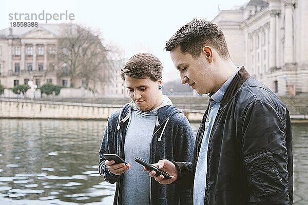 Zwei asoziale handyabhängige männliche Teenager stehen zusammen und benutzen ein Smartphone  Technologiekonzept  städtische Uferlage in Berlin Deutschland