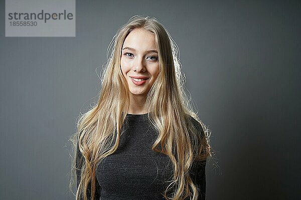 Teenager-Mädchen mit langen blonden Haaren und strahlendem Lächeln - Studio-Porträt einer jungen Frau vor grauem Hintergrund mit Kopierraum