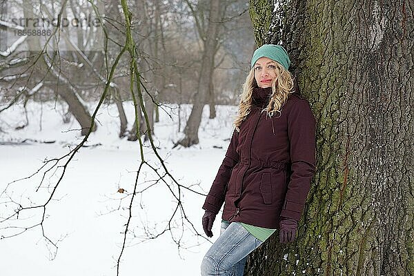 Blonde Frau lehnt an einem Baum in einem verschneiten Park im Winter