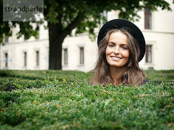 Glücklich lächelnde junge Frau in einem Park mit Blick über eine Hecke