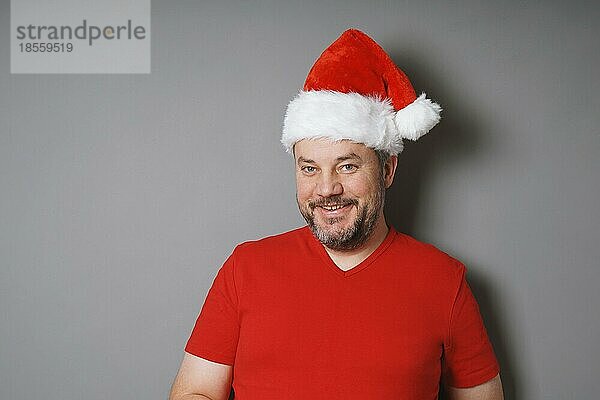 Lächelnder Mann mittleren Alters mit ergrauendem Bart  der eine Weihnachtsmannmütze und ein rotes T-Shirt trägt - Konzept für echte Menschen zu Weihnachten