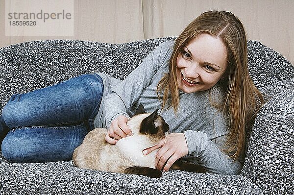 Glückliche junge Frau entspannt sich auf der Couch mit ihrer Siamkatze
