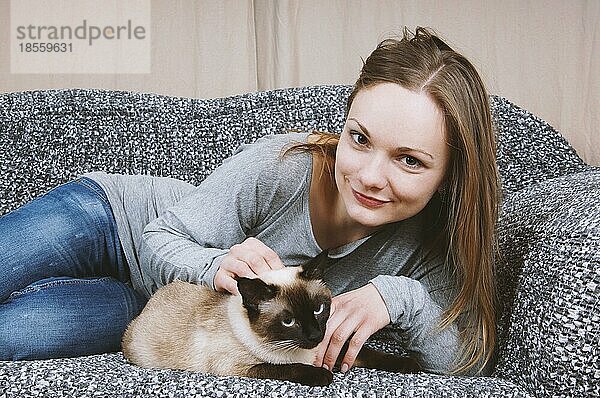 Glückliche junge Frau entspannt sich auf dem Sofa mit ihrer Siamkatze