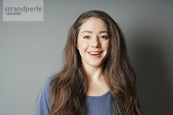 Glückliche junge Frau in den 20ern mit natürlichem Make-up und brünetten langen Haaren lächelt zähnefletschend - vor grauem Hintergrund mit Copy Space